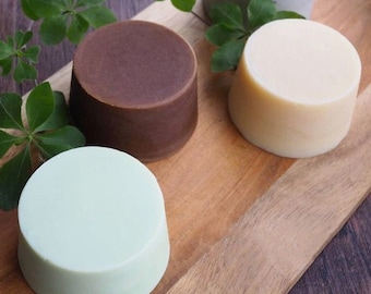 May include: Three round bars of soap on a wooden surface. The soap bars are brown, white, and light green.