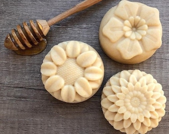 May include: Three round, light-coloured soaps with floral designs, including a sunflower, a daisy, and a layered flower. A wooden honey dipper rests beside the soaps, with a small amount of honey visible. The soaps are arranged on a wooden surface.