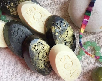 May include: A close-up of a pile of handmade soap bars. The bars are oval-shaped and have a black and gold design. Some of the bars have the word "SOAP" embossed on them.