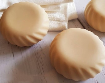 May include: Three round, beige-coloured soap bars with a textured, rippled surface. The soap bars are arranged on a white wooden surface.