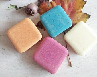 May include: Four square-shaped solid shampoo bars in different colors: light orange, light blue, light pink, and white. The bars are arranged on a white surface with a dried autumn leaf.