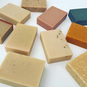 Peut inclure: Assortiment de savons artisanaux de différentes couleurs et textures, notamment beige, marron, orange, rose et vert. Les savons sont disposés sur une surface blanche.