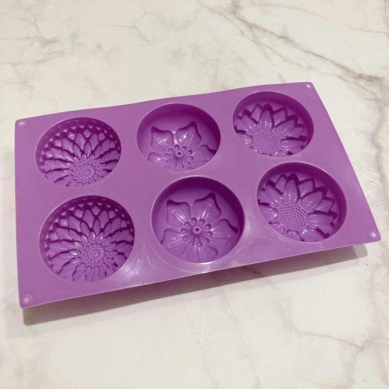 May include: A lavender silicone mold with six circular cavities, each featuring a floral design. The mold includes three sunflower and three other flower patterns, perfect for crafting soaps or bath bombs.