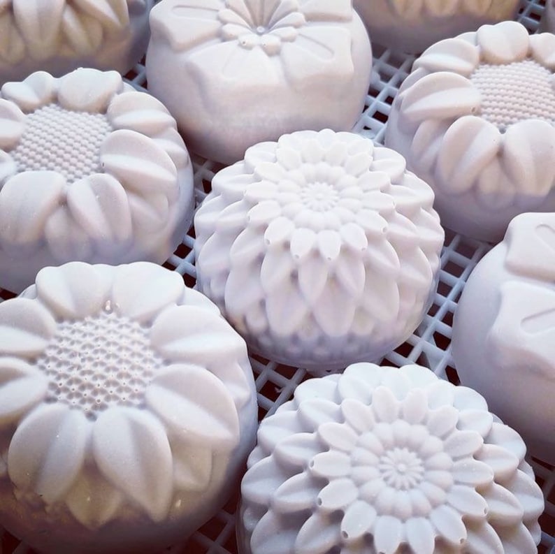 May include: A collection of white, floral-shaped soaps. The soaps are round with detailed petal and center designs, resembling sunflowers and other blossoms. They are arranged on a white, grid-like surface, creating a visually appealing display.