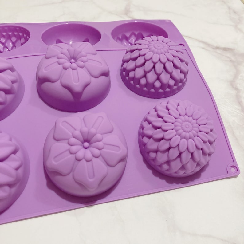 May include: A lavender silicone mold with six cavities, each designed with a floral pattern. The mold features three different flower designs, including a detailed chrysanthemum and two other stylized floral shapes. The mold is likely for making soaps, candles, or other crafts.