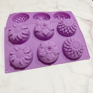 May include: A lavender silicone mold with six cavities. The mold features three different floral designs: sunflowers, a flower with rounded petals, and a flower with layered petals. The mold is designed for making soaps, candles, or other crafts.