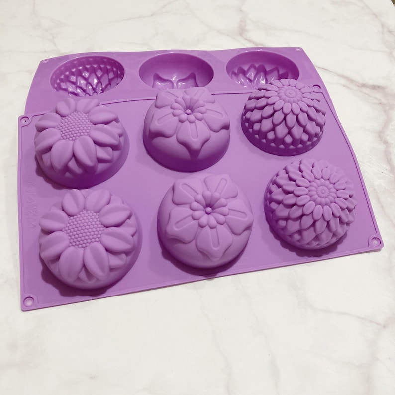May include: A lavender silicone mold with six cavities. The mold features three different floral designs: sunflowers, a flower with rounded petals, and a flower with layered petals. The mold is designed for making soaps, candles, or other crafts.