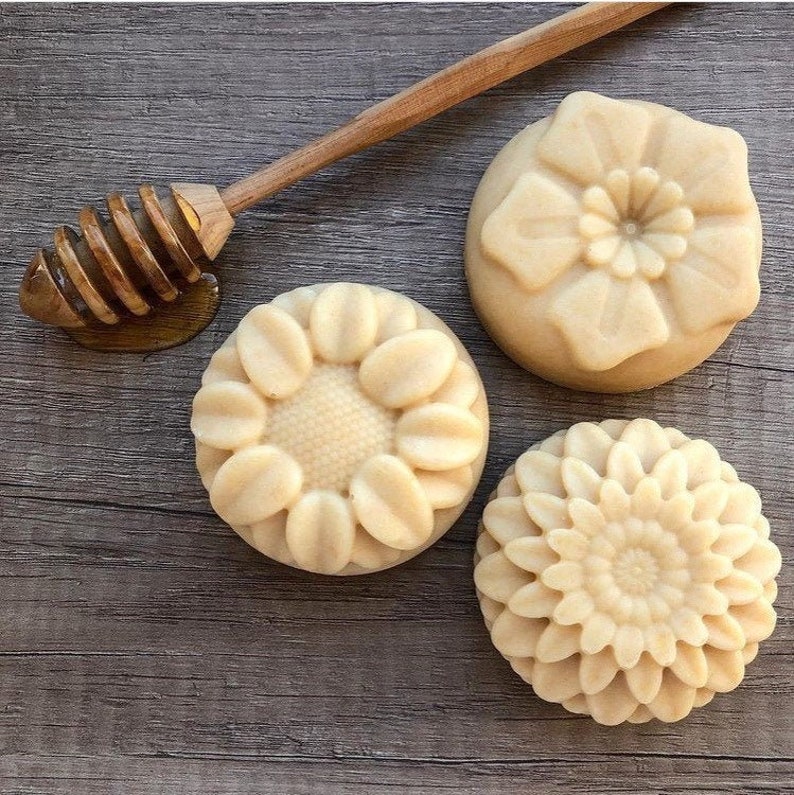 May include: Three round, light-colored soaps with floral designs, including a sunflower, a daisy, and a layered flower. A wooden honey dipper rests beside the soaps, with a small amount of honey visible. The soaps are arranged on a wooden surface.