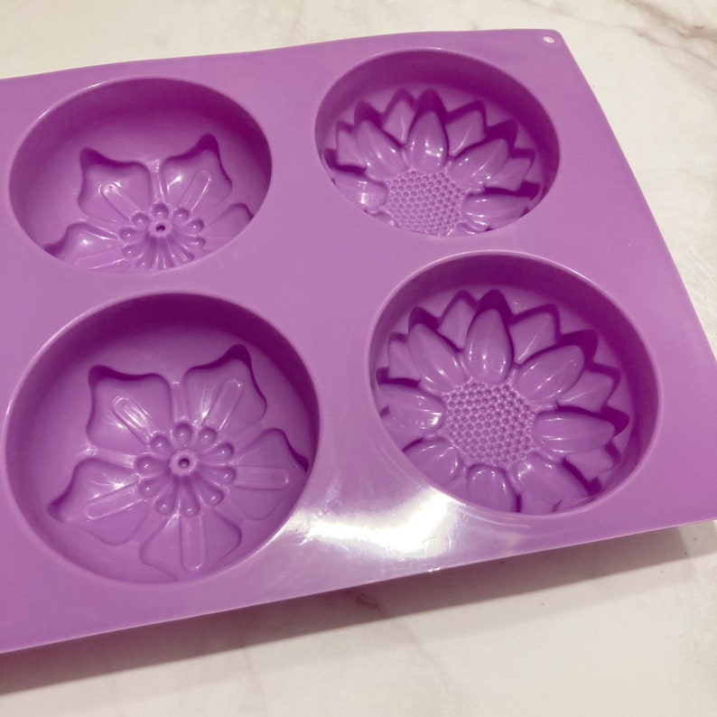 May include: A lavender silicone mold with four circular cavities, each featuring a floral design. Two cavities have a sunflower pattern, and the other two have a different flower design. The mold is designed for crafting soaps or other similar items.