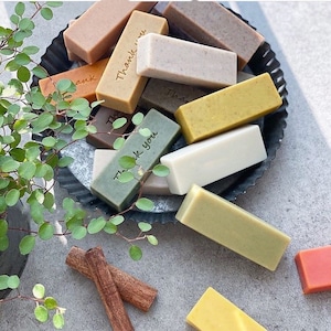 May include: A collection of rectangular bars of soap in various colours, including green, brown, beige, and yellow, arranged on a black metal tray. Some of the bars have the words "Thank you" and "This" inscribed on them. Two cinnamon sticks are on a grey surface.