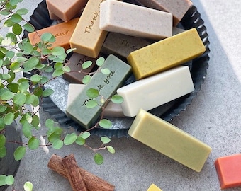 Puede incluir: Una colección de barras de jabón rectangulares de varios colores, incluyendo verde, marrón, beige y amarillo, dispuestas en una bandeja de metal negro. Algunas de las barras tienen las palabras "Gracias" y "Esto" inscritas en ellas. Dos palitos de canela están sobre una superficie gris.