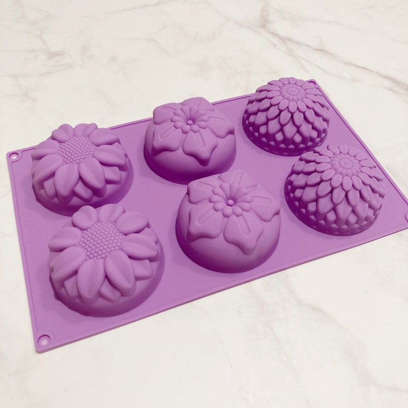 May include: A lavender silicone mold with six floral-shaped cavities. The mold features three sunflower designs and three other flower designs. The cavities are arranged in two rows of three. The mold is designed for making soaps, candles, or other crafts.