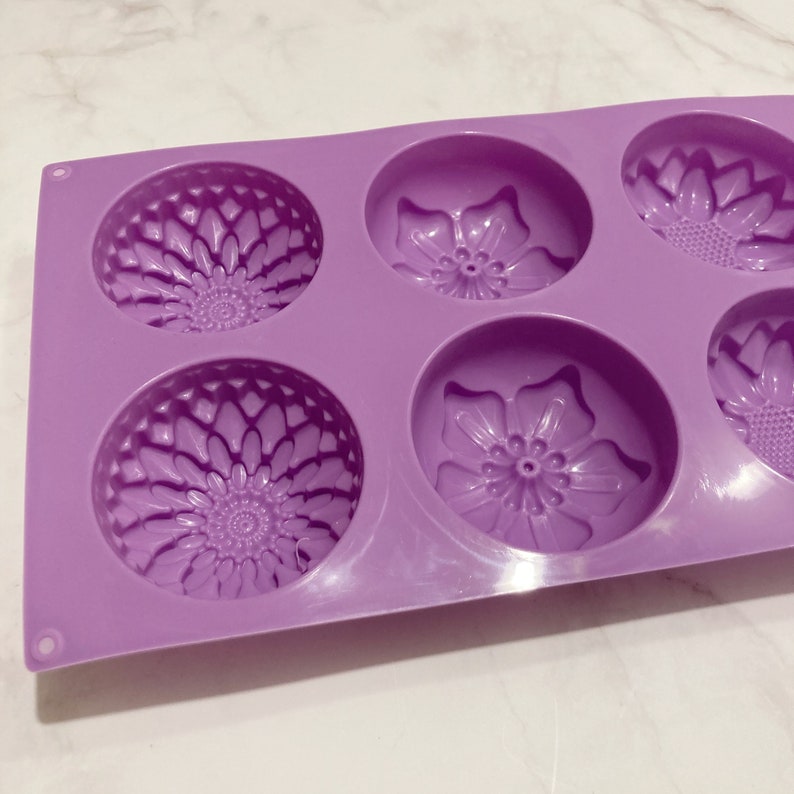 May include: A lavender silicone mold with six circular cavities, each featuring a floral design. The mold has three cavities with a sunflower pattern and three with a flower pattern. The mold is designed for crafting soaps, candles, or other DIY projects.