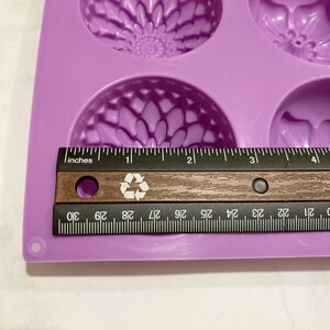 May include: A lavender silicone mold with floral designs, including a sunflower and other flower shapes. A wooden ruler with black markings is placed across the mold, measuring approximately 4 inches in length.