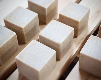 May include: A set of eight square-shaped bars of soap, arranged in rows on a wooden surface. The soap is a light beige color with a subtle shimmer.