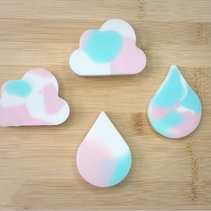Pode incluir: Quatro barras de sabão feitas à mão com a forma de nuvens e gotas de chuva. Os sabões são brancos com redemoinhos rosa e azuis.