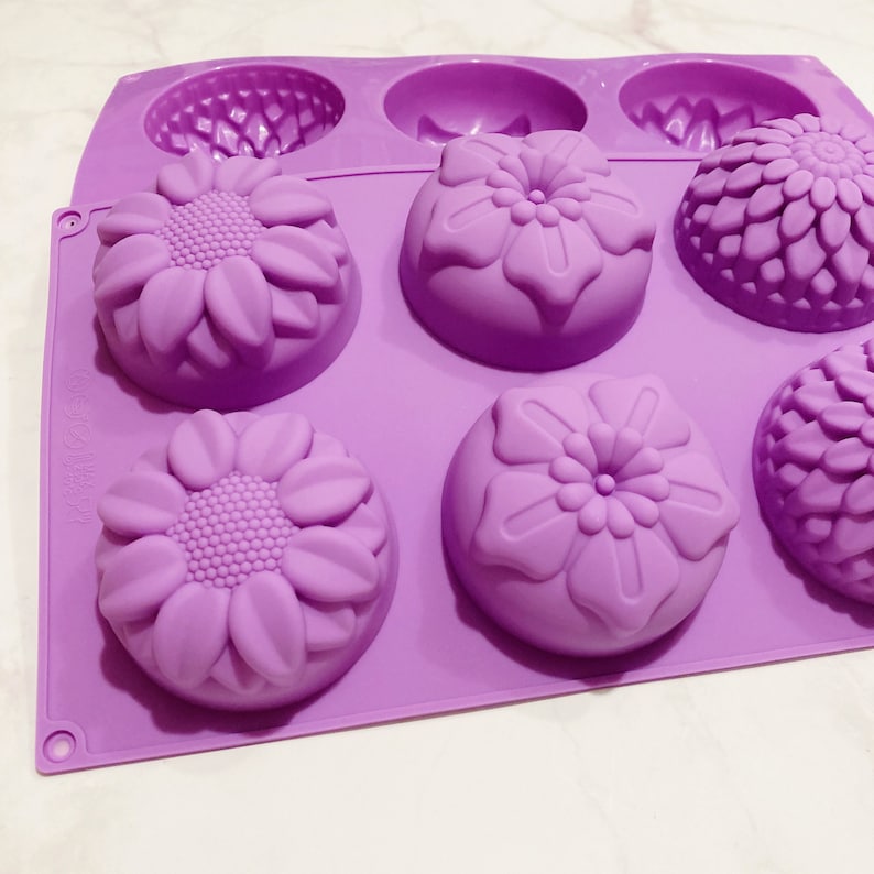 May include: A lavender silicone mold with six floral designs. The mold features sunflower, daisy, and other flower shapes. The mold is designed for making soaps, candles, or other crafts. The mold is made of flexible silicone.