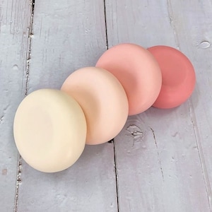May include: Four circular objects in a row, each a different shade of pink and cream. The colours range from a pale cream to a deep rose. The objects are on a light grey wooden surface.