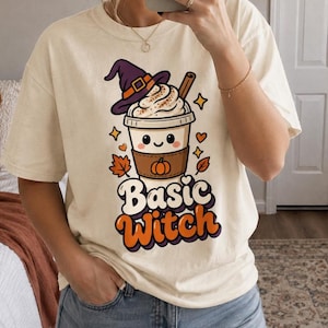 Op de afbeelding: Een crèmekleurig T-shirt met korte mouwen en een afbeelding van een koffiebeker met een lachend gezicht, die een paarse heksenhoed draagt. De beker heeft een bruine hoes met een pompoenicoon. De tekst Basic Witch staat in oranje en wit eronder.