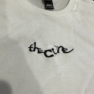 Pode incluir: Camiseta branca com um logotipo "The Cure" bordado em preto. A camiseta é feita por "Dist. Rick" e é tamanho S.