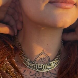 May include: A gold-toned necklace with a geometric design is worn around the neck. The necklace is paired with a brown scarf with sequin details. The person has red hair and a nose ring.