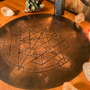 May include: A round, bronze-colored metal plate with an etched geometric design. The plate is surrounded by clear quartz crystals and other decorative stones. The plate's design features intersecting lines and circular patterns, creating a complex, symmetrical pattern.