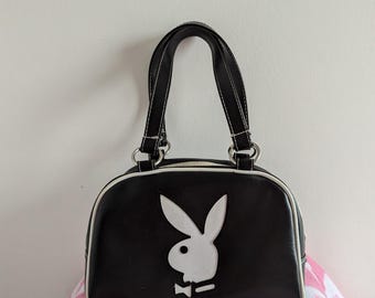 Peut inclure: Un sac à main en similicuir noir avec un logo de lapin Playboy blanc. Le sac a une poignée supérieure et une forme en dôme.