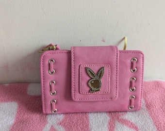 May include: A rectangular pink wallet featuring a central flap with a metallic gold-toned rabbit logo emblem. The wallet includes silver-toned metal grommets along the left and right sides, with a zipper closure visible on the top left corner.