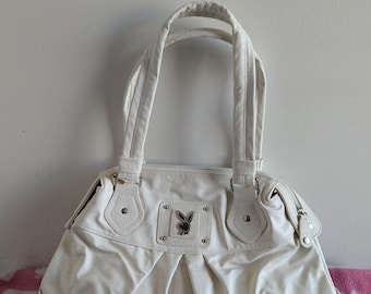 May include: Cream-colored handbag with a pleated front and two top handles. The bag features a silver-toned metal emblem with a bunny logo. The bag is sitting on a pink and white patterned surface.