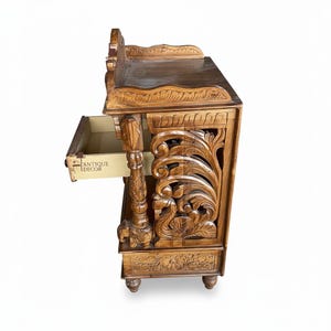 May include: A brown wooden side table featuring intricate hand-carved floral and swan motifs on the side panels. The table includes a single pull-out drawer with the text ANTIQUE DECOR printed on the side, supported by carved pillar legs.