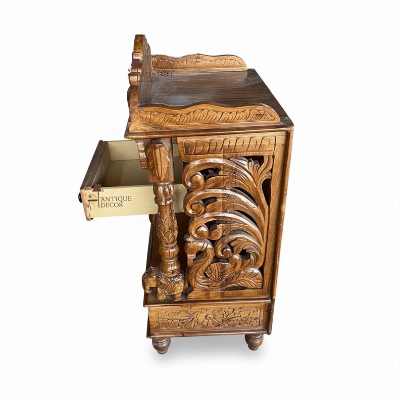 May include: A brown wooden side table featuring intricate hand-carved floral and swan motifs on the side panels. The table includes a single pull-out drawer with the text ANTIQUE DECOR printed on the side, supported by carved pillar legs.