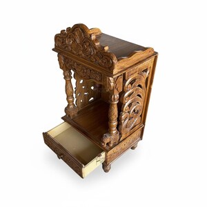 May include: A brown wooden temple with intricate carvings, featuring a flat top, two front pillars with elephant-shaped bases, and a pull-out drawer at the bottom. The structure has detailed floral and scrollwork patterns on the sides and top arch.