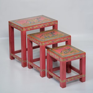 May include: A set of three nesting tables in varying sizes, each featuring a red wooden frame and a rectangular top. The tops are painted with a central elephant illustration in grey and gold, surrounded by a green floral border on a red background.