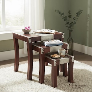 May include: A set of three nesting tables crafted from dark brown wood, each featuring a central inlay of textured light gray fabric with a decorative white lace border. The tables are arranged in a cascading formation, displaying a smooth finish.