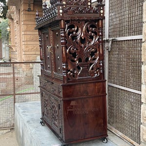 K&ouml;nnte beinhalten: Ein dunkelbrauner Holzschrank mit aufwendigen Schnitzereien und Zierdetails. Der Schrank verf&uuml;gt &uuml;ber mehrere T&uuml;ren, Schubladen und dekorative Elemente, darunter Blumenmuster und eine kronenartige Spitze. Der Schrank steht auf Rollen und tr&auml;gt den Text "VM ANTIQUE DECOR".