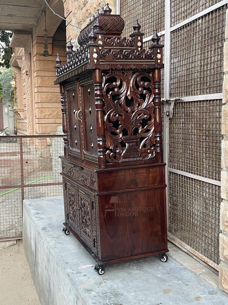 K&ouml;nnte beinhalten: Ein dunkelbrauner Holzschrank mit aufwendigen Schnitzereien und Zierdetails. Der Schrank verf&uuml;gt &uuml;ber mehrere T&uuml;ren, Schubladen und dekorative Elemente, darunter Blumenmuster und eine kronenartige Spitze. Der Schrank steht auf Rollen und tr&auml;gt den Text "VM ANTIQUE DECOR".