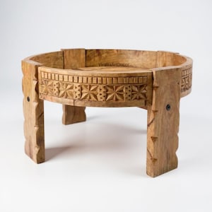 May include: A round wooden pedestal stand with four thick legs, featuring a carved star pattern along the outer rim and the text VirattaArtDecor visible on the inner surface. The light brown wood has a natural grain and dark metal hardware.