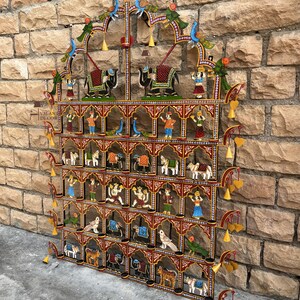 Puede incluir: Un adorno de pared rectangular de metal con varios niveles, que presenta un marco rojo con patrones florales blancos. Contiene numerosos estantes peque&ntilde;os que exhiben figuras pintadas de animales, aves y personas, con dijes amarillos.