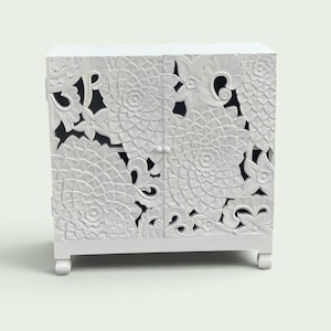 Könnte beinhalten: Ein weißer Schrank mit zwei Türen, die mit detaillierten, tiefen Blumenreliefs verziert sind. Das Design zeigt große, geschichtete Blütenblattmuster und geschwungene organische Formen auf der Vorderseite, getragen von vier kleinen, abgerundeten Füßen.