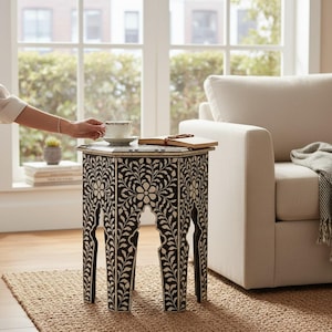 May include: A black and white hexagonal side table with a floral inlay design. The table has arched cutouts on the sides. A cup and saucer sit on top, next to an open book. The table is on a woven rug.