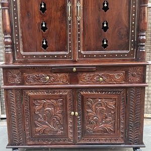 K&ouml;nnte beinhalten: Ein dunkelbrauner Holzschrank mit aufwendigen Schnitzereien und Messingbeschl&auml;gen. Der Schrank hat zwei T&uuml;ren mit floralen Schnitzereien, zwei Schubladen und einen oberen Bereich mit glockenf&ouml;rmigen Ausschnitten. Der Schrank steht auf Rollen.