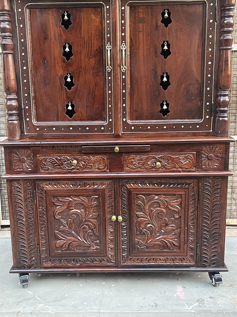 K&ouml;nnte beinhalten: Ein dunkelbrauner Holzschrank mit aufwendigen Schnitzereien und Messingbeschl&auml;gen. Der Schrank hat zwei T&uuml;ren mit floralen Schnitzereien, zwei Schubladen und einen oberen Bereich mit glockenf&ouml;rmigen Ausschnitten. Der Schrank steht auf Rollen.