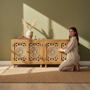 May include: A light brown wooden sideboard featuring four cabinet doors with intricate circular floral carvings. The piece stands on short legs and has a rectangular top surface. The wood grain is visible throughout the structure of the furniture.