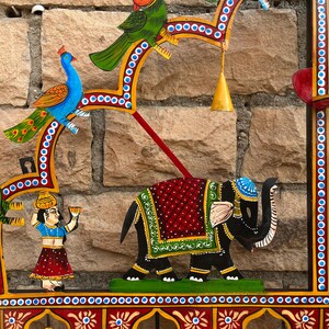 Puede incluir: Un colgante de pared decorativo con figuras pintadas a mano de un elefante, p&aacute;jaros y personas. La pieza incluye un elefante negro central con una manta roja y verde, un pavo real azul, un loro verde y figuras que llevan recipientes.