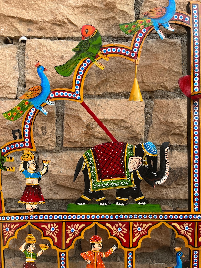 Puede incluir: Un colgante de pared decorativo con figuras pintadas a mano de un elefante, p&aacute;jaros y personas. La pieza incluye un elefante negro central con una manta roja y verde, un pavo real azul, un loro verde y figuras que llevan recipientes.