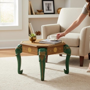 Könnte beinhalten: Ein sechseckiger Beistelltisch aus Holz mit einer naturfarbenen Tischplatte und drei Beinen, die als grüne Elefantenköpfe mit goldenen Akzenten geschnitzt sind. Die Seiten sind mit goldenen Blumenmotiven und einer kleinen Sukkulente verziert.