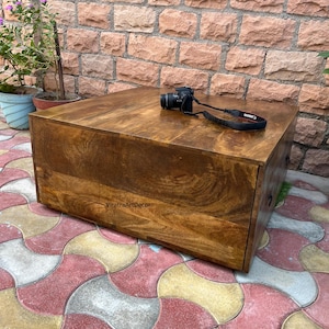 Könnte beinhalten: Ein quadratischer, niedriger Couchtisch aus massivem braunem Holz mit sichtbarer Maserung. Auf der Oberfläche ist der Text ViratraArtDecor in Schwarz aufgedruckt. Eine schwarze Digitalkamera mit Trageriemen liegt auf dem Tisch.
