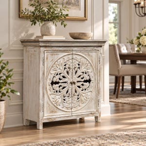 Puede incluir: Un gabinete rectangular de madera con un acabado blanco desgastado, que presenta dos puertas adornadas con una gran y compleja talla floral circular. El mueble tiene una superficie superior plana, una base resistente y pequeños pomos de metal oscuro.