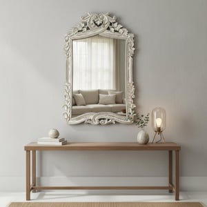 Puede incluir: Un espejo de pared rectangular con un marco de madera blanca desgastada y adornos tallados de hojas y volutas. El espejo está montado sobre una mesa consola de madera larga y estrecha que sostiene un pequeño jarrón y una lámpara de mesa.