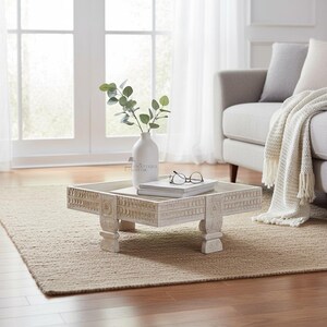 Könnte beinhalten: Ein quadratischer, flacher Couchtisch aus Holz mit weißem Antik-Finish und detaillierten Schnitzereien an den Kanten und Beinen. Auf der Tischplatte stehen eine weiße Keramikvase mit grünem Blattwerk sowie ein Stapel Bücher mit Brille.
